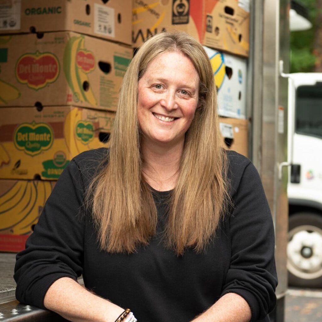 A person with long hair, wearing a black shirt, leans against a truck filled with boxes of fruit.