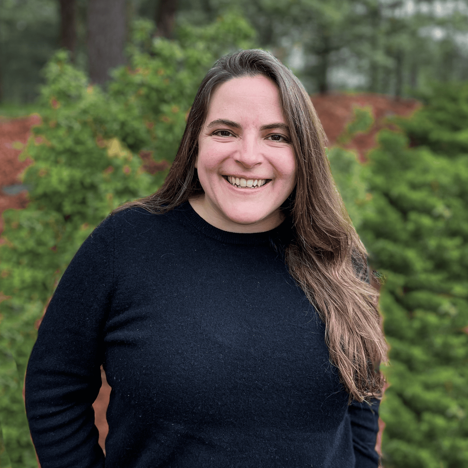 A woman with long brown hair smiles outdoors, standing in front of green foliage while wearing a black sweater.