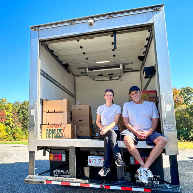 Two people sitting in the back of a moving truck with boxes of food donated to Boston area food recovery organization Lovin Spoonfuls.