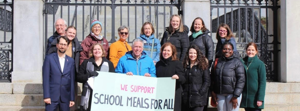 A group of people working as advocates for school lunch programs standing in front of the Massachusetts State House holding a sign.