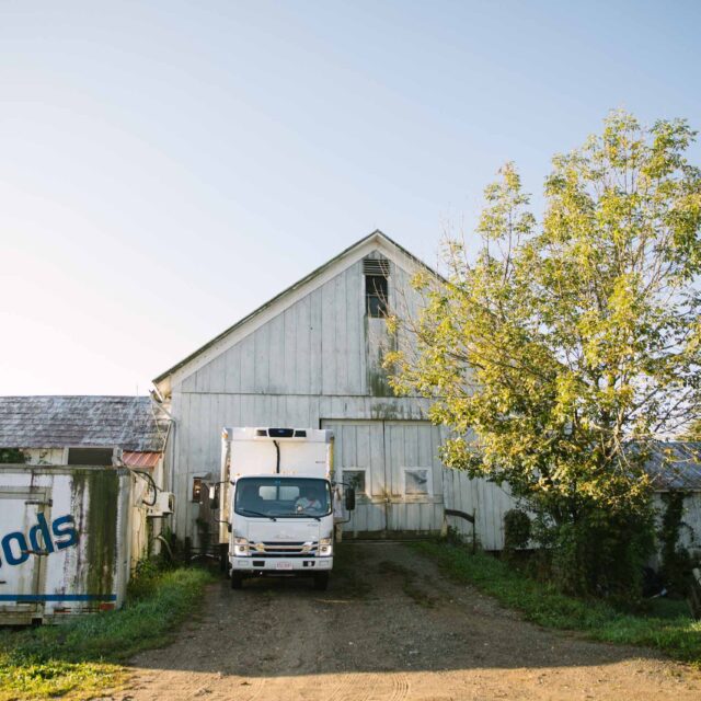 A truck is parked in front of a white barn on a farm while donated food is being picked up by Boston area food recovery charity Lovin Spoonfuls.