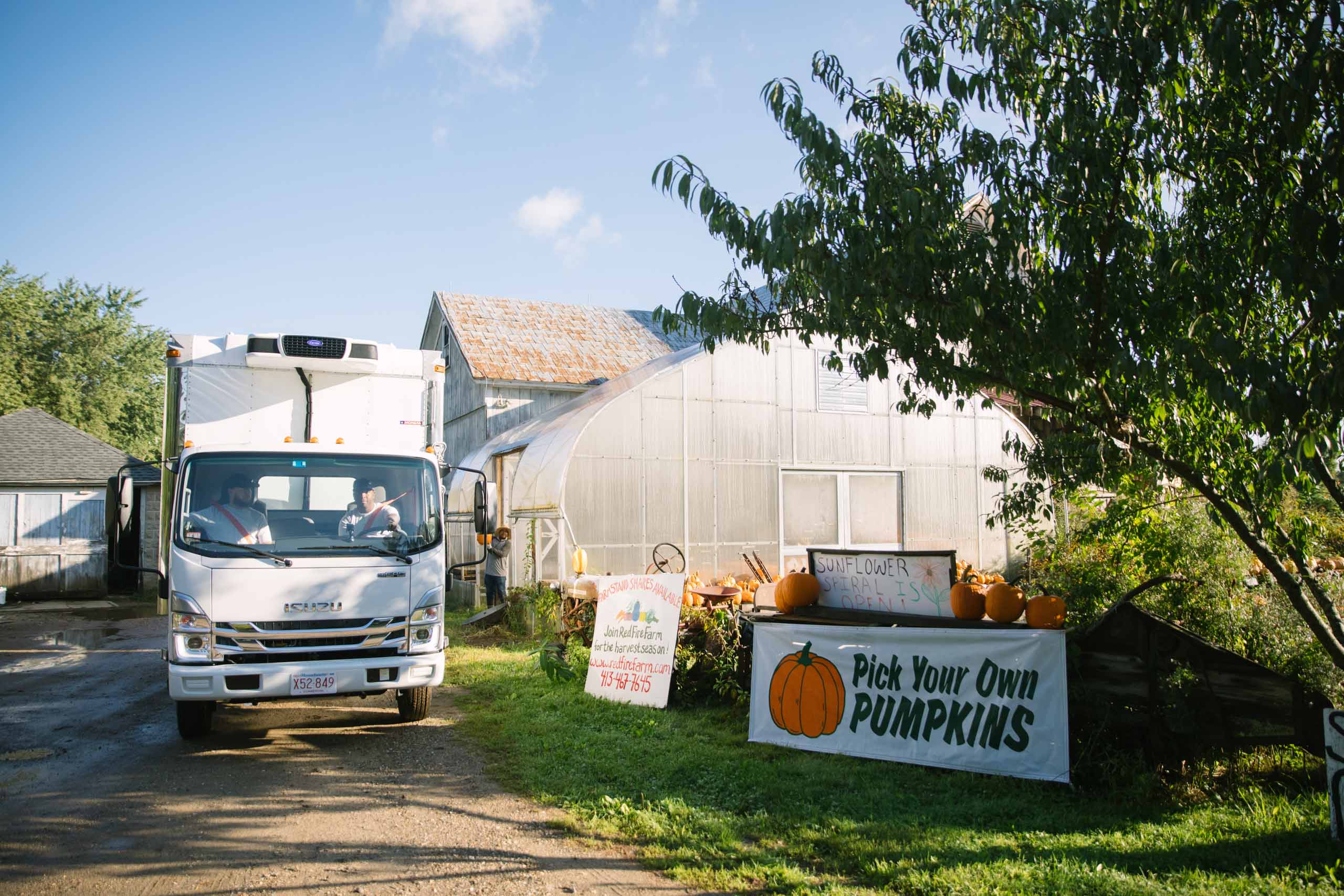 Two men in a white truck parked on a dirt road at a farm picking up pumpkins donated to Boston area food recovery organization Lovin Spoonfuls.