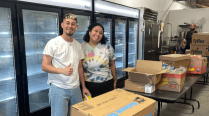 Two people stand in front of a row of freezers, smiling at the camera, surrounded by open boxes and packaged goods. One person gives a thumbs-up.