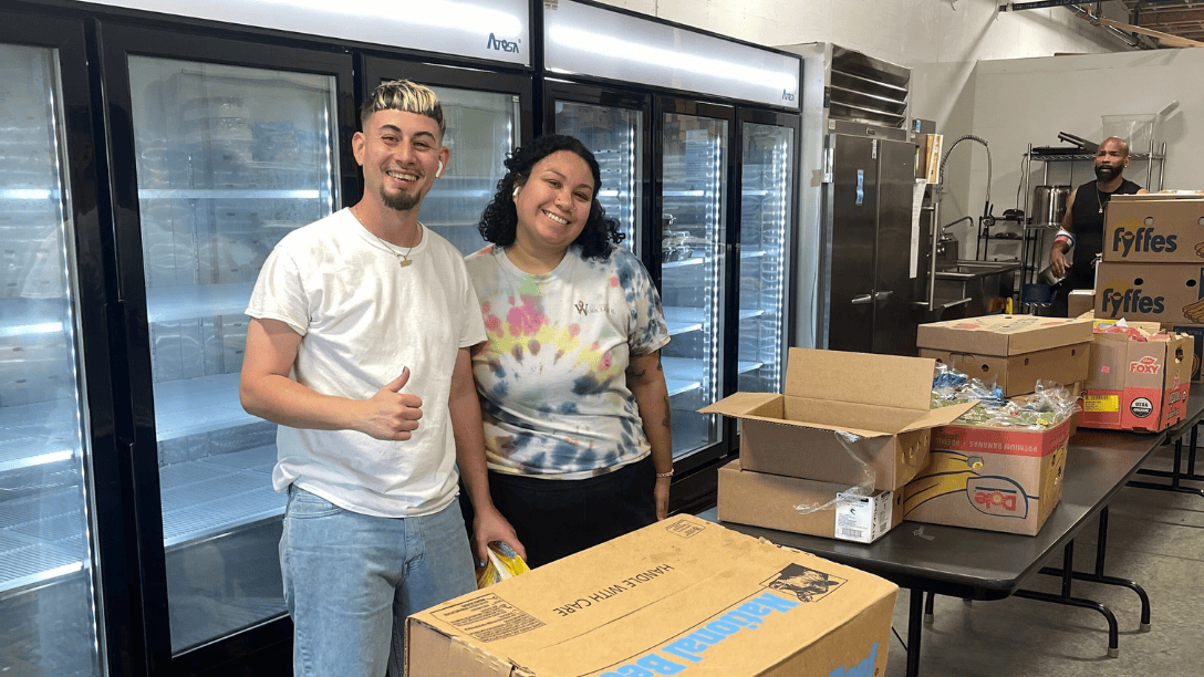 Two people stand in front of a row of freezers, smiling at the camera, surrounded by open boxes and packaged goods. One person gives a thumbs-up.