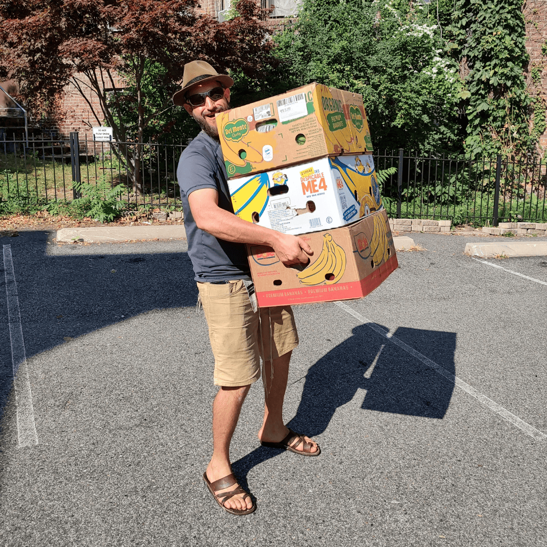 A man wearing a hat and sunglasses carries three large cardboard boxes in the sunlit parking area.