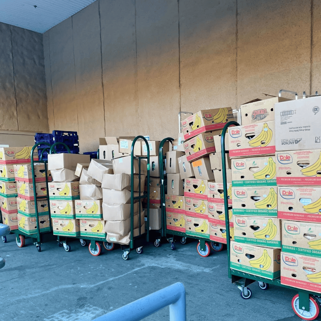 Carts loaded with cardboard boxes and paper bags, filled with bananas and other items, are lined up outside a building.