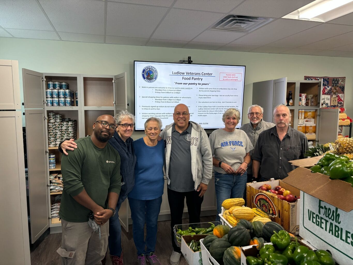 A group of people standing in a food pantry with fresh vegetables and canned goods on display. There's a screen in the background showing information about the Ludlow Veterans Center Food Pantry.