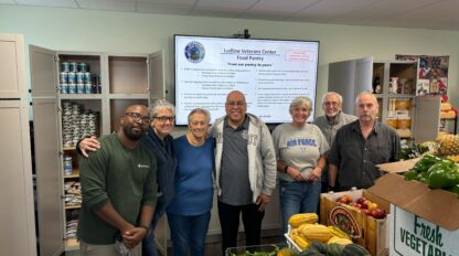 A group of people standing in a food pantry with fresh vegetables and canned goods on display. There's a screen in the background showing information about the Ludlow Veterans Center Food Pantry.