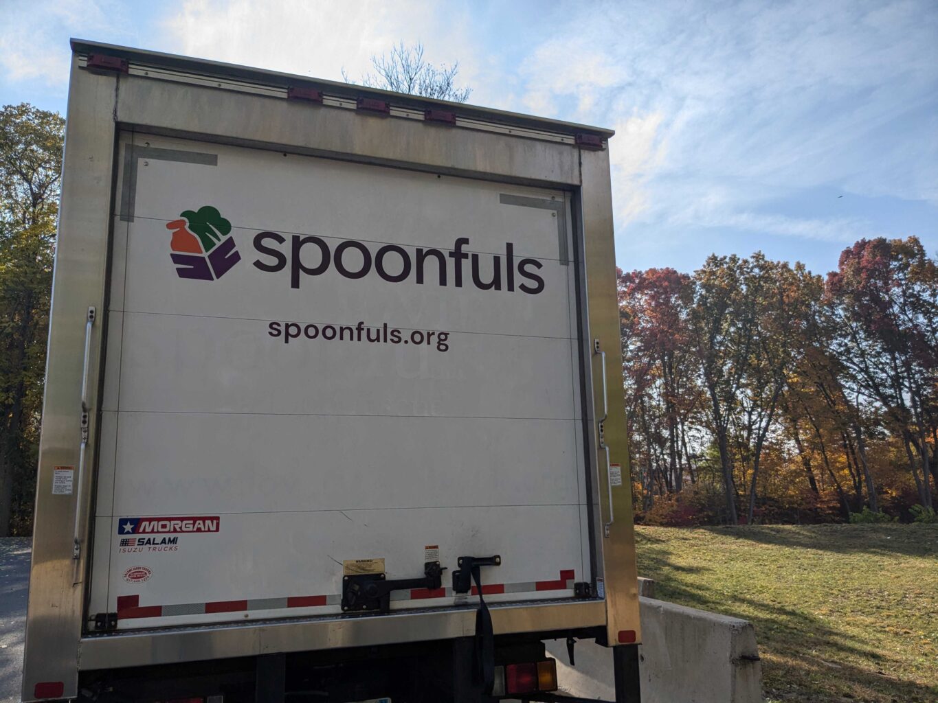 The back of a delivery truck displaying the logo and website of Spoonfuls, with trees and a clear sky in the background.