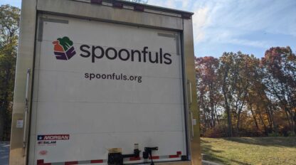 The back of a delivery truck displaying the logo and website of Spoonfuls, with trees and a clear sky in the background.