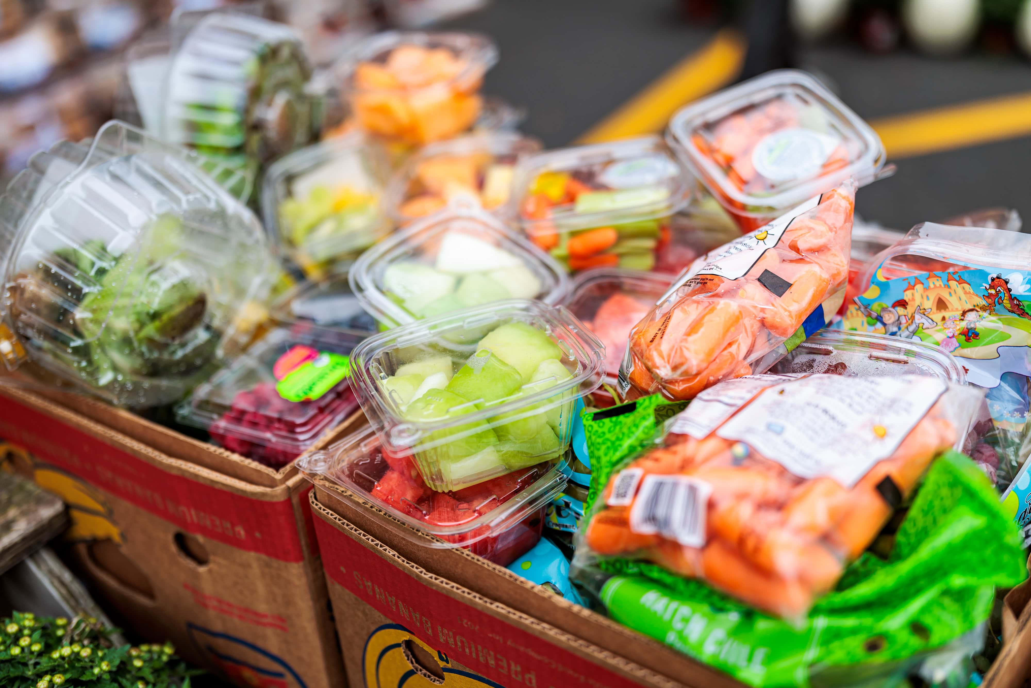 A variety of packaged fresh fruits and vegetables are displayed on cardboard boxes at an outdoor market.