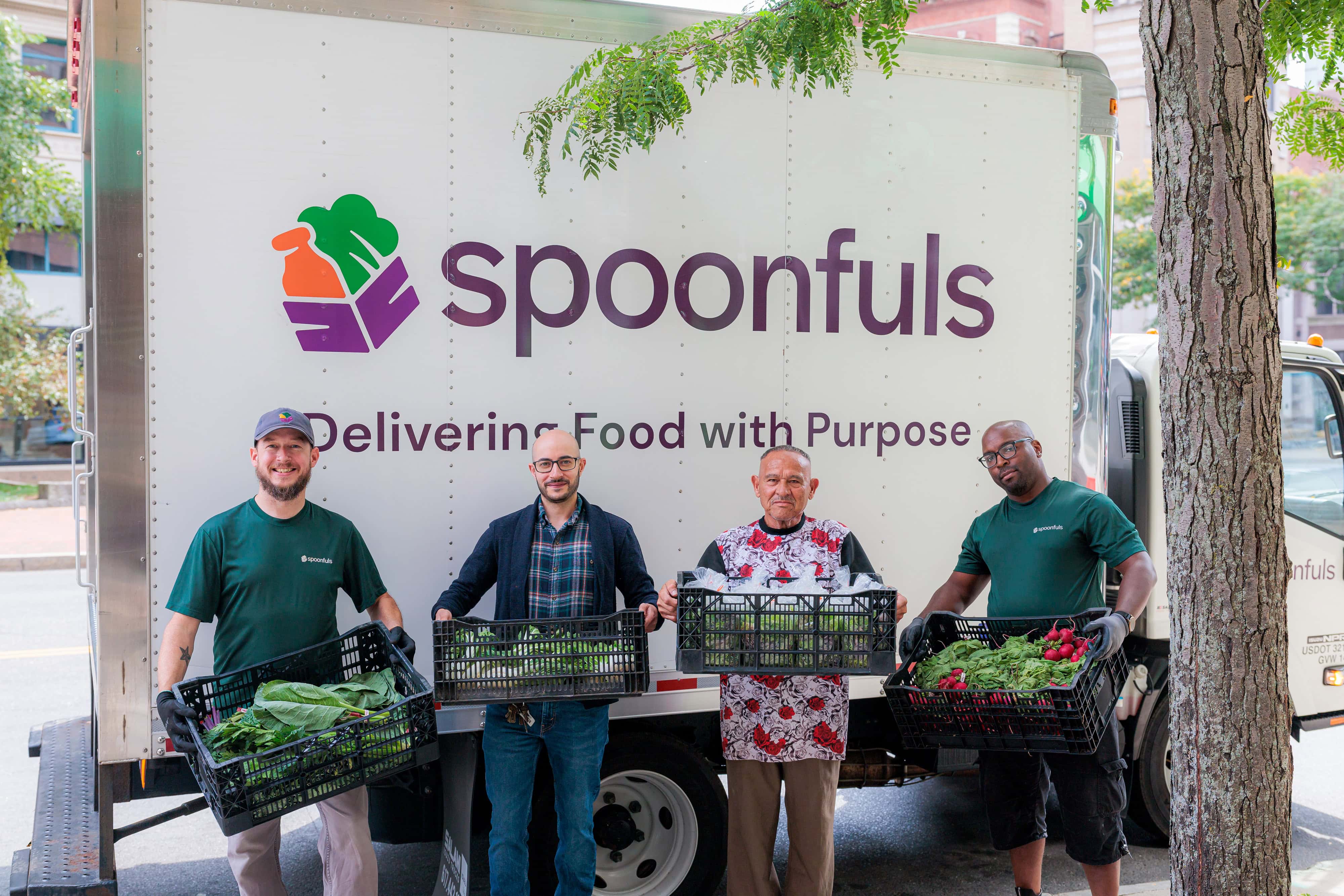 Four people stand in front of a "Spoonfuls" truck, each holding crates of fresh produce.