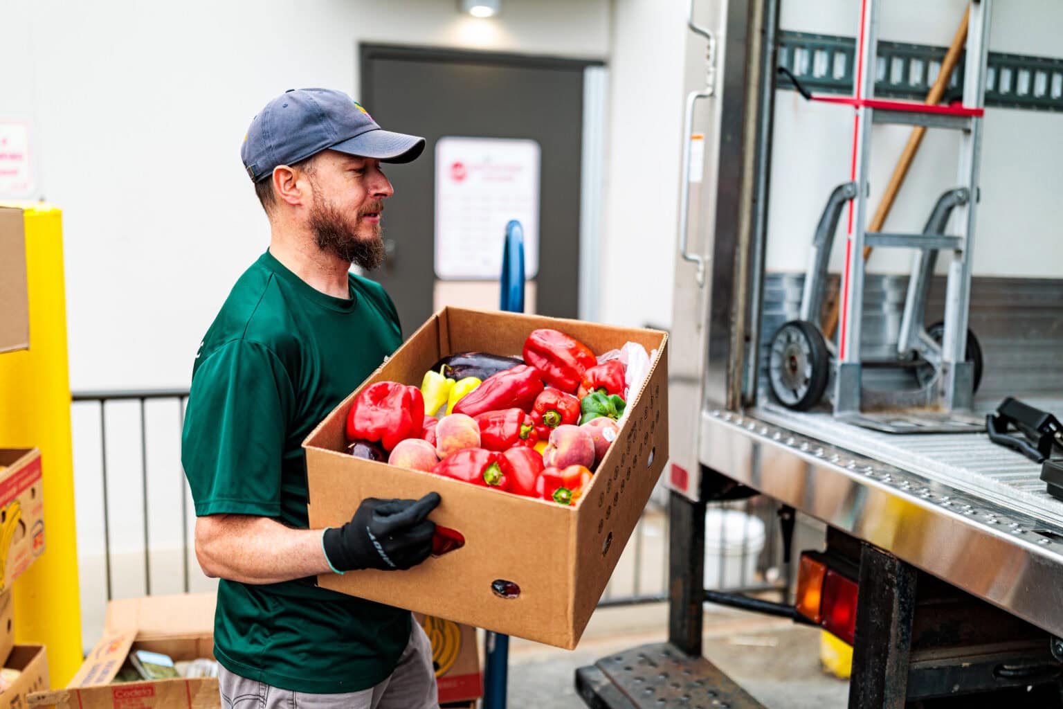 A man in a green shirt and cap carries a box of assorted vegetables to a truck.