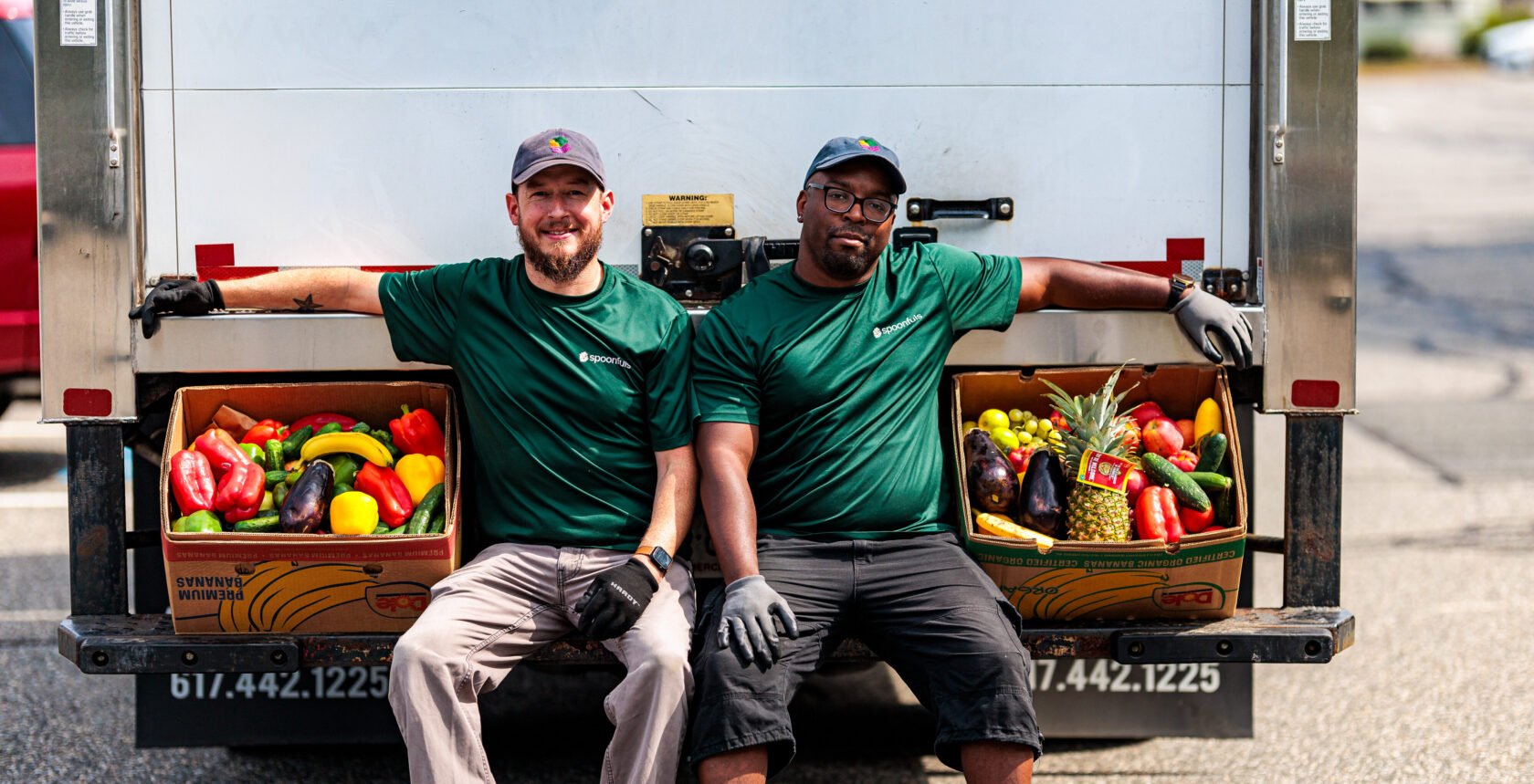 Two men in green shirts sit on a truck bumper with arms resting on boxes of assorted fruits and vegetables.