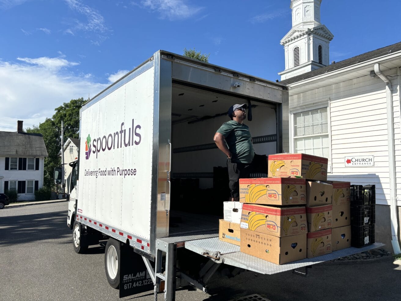 A man stands in the back of a Spoonfuls delivery truck, which is parked next to a white building. Several boxes of bananas are stacked inside.