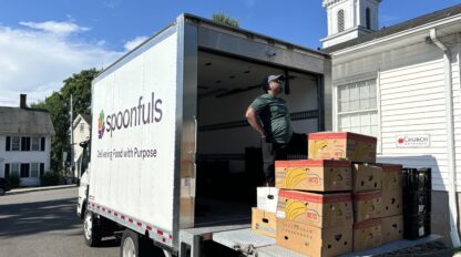 A man stands in the back of a Spoonfuls delivery truck, which is parked next to a white building. Several boxes of bananas are stacked inside.