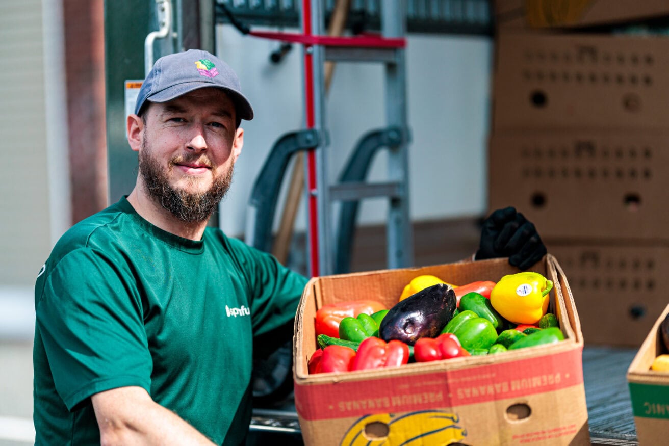 A person in a green shirt and cap holds a box of assorted vegetables, including bell peppers and an eggplant, beside a vehicle loaded with more boxes.