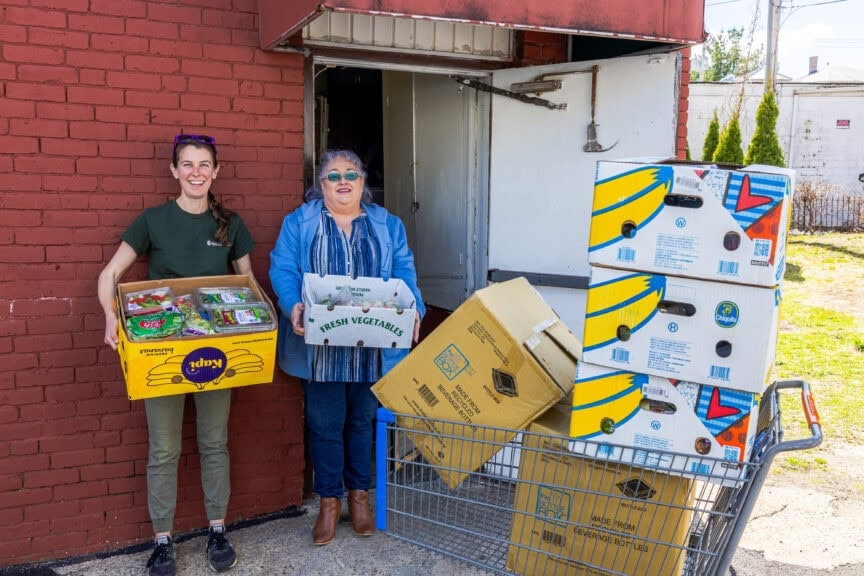 Two people stand beside a brick building, holding boxes of food near a cart stacked with cardboard boxes.