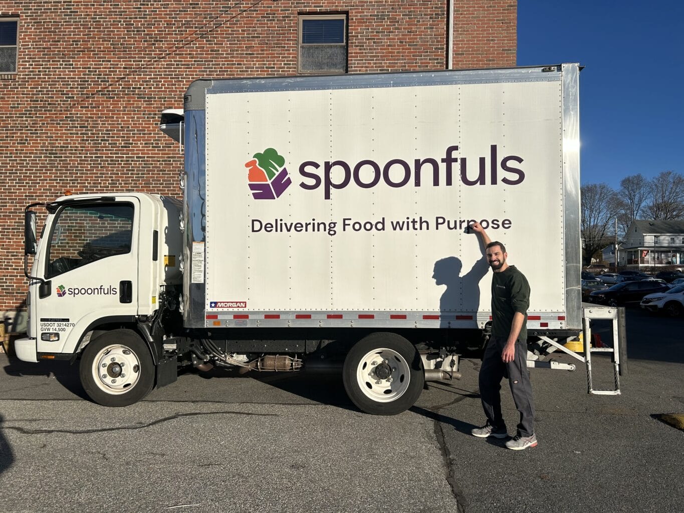 A man stands beside a Spoonfuls delivery truck with the slogan "Delivering Food with Purpose" on the side.