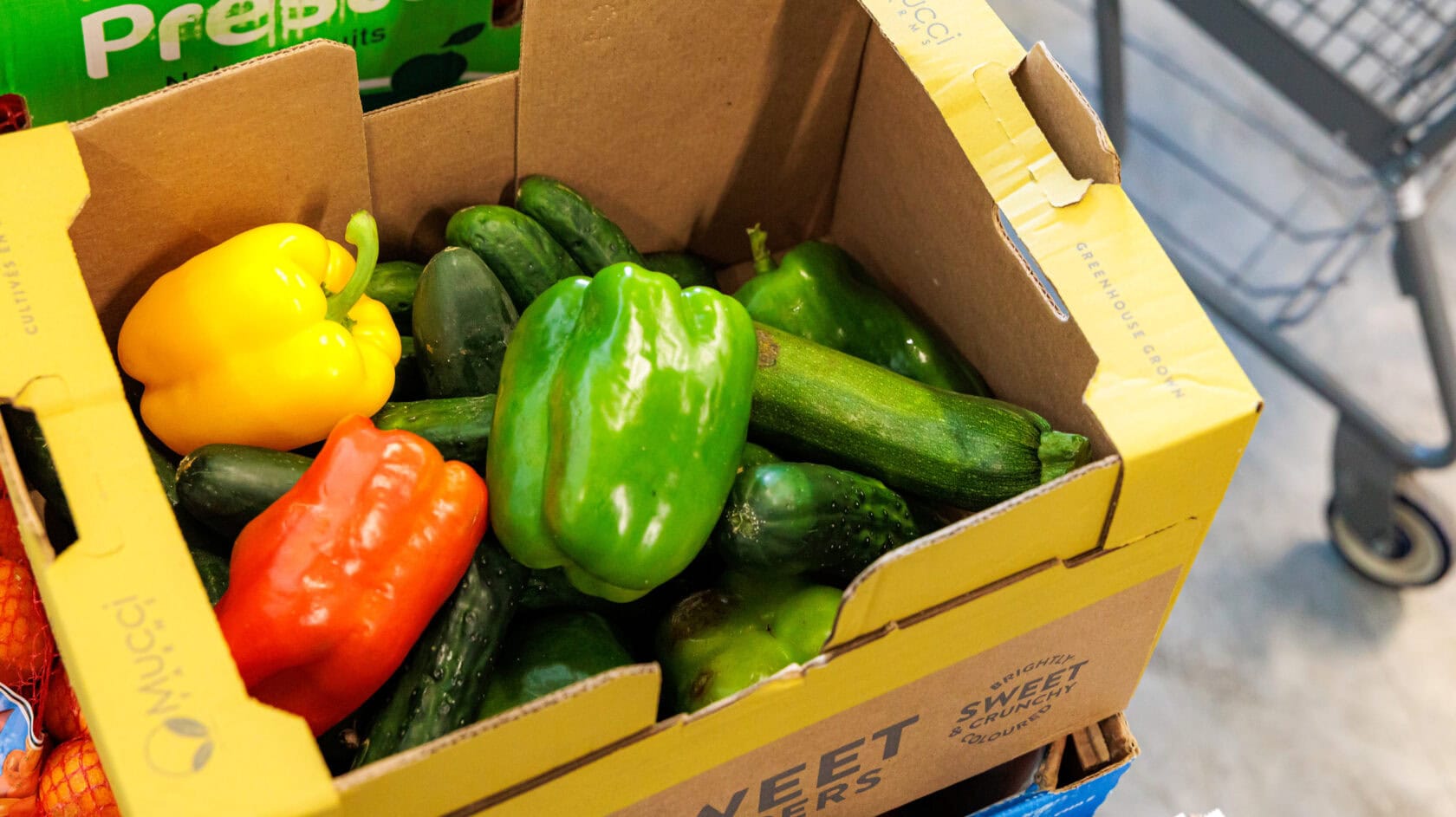 A cardboard box holds green, yellow, and red bell peppers with several cucumbers, setting the scene for a fresh market story. A shopping cart is visible in the background.