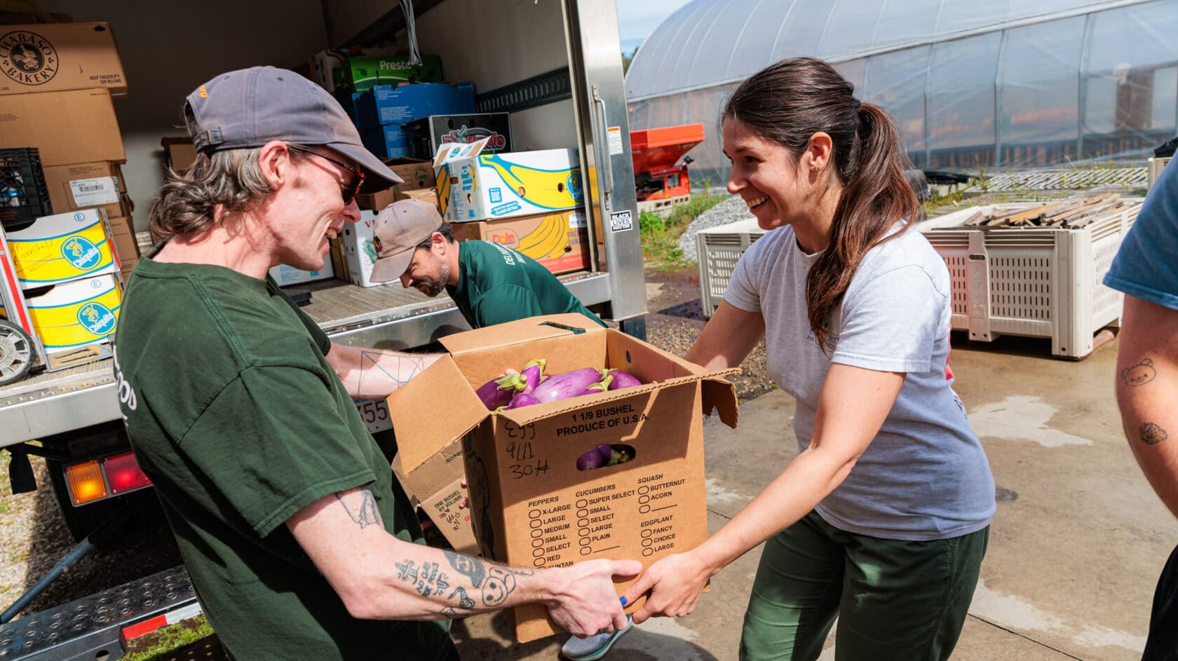 Two people smiling as they work together to unload a box of produce from a truck near a greenhouse, capturing the story of teamwork and fresh beginnings.