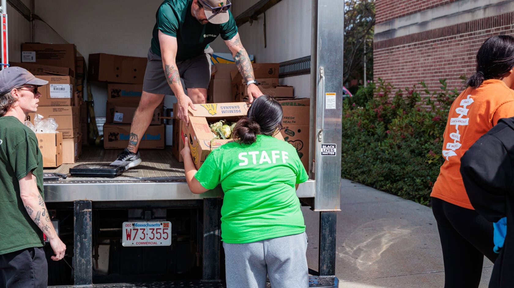 People unload boxes of produce from a truck, with one person wearing a "STAFF" shirt and others assisting near the vehicle in a sunny outdoor setting.
