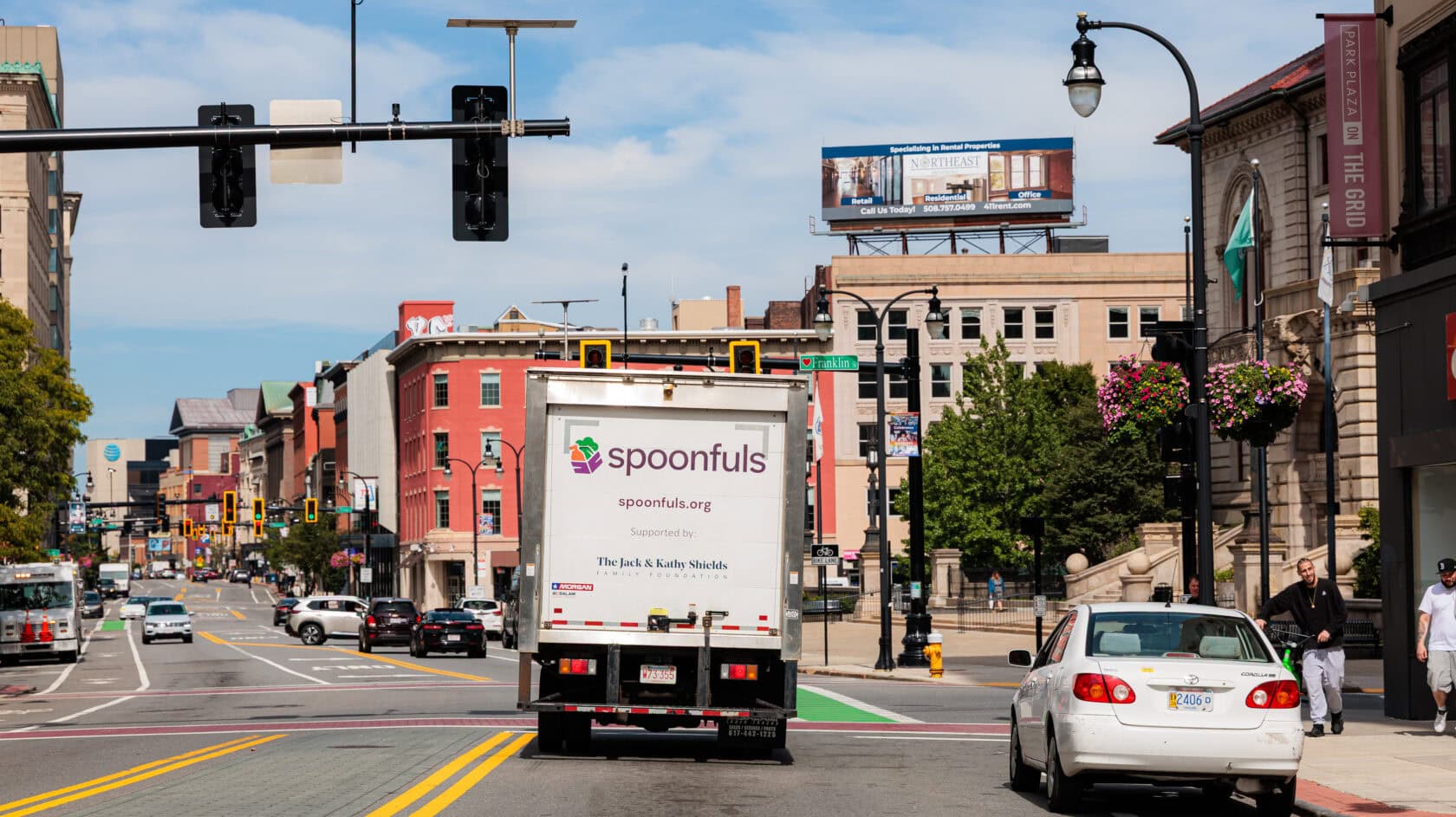 A Spoonfuls delivery truck drives down a city street lined with brick buildings, traffic lights, and cars, telling a story of community as it rolls past a visible billboard in the background.