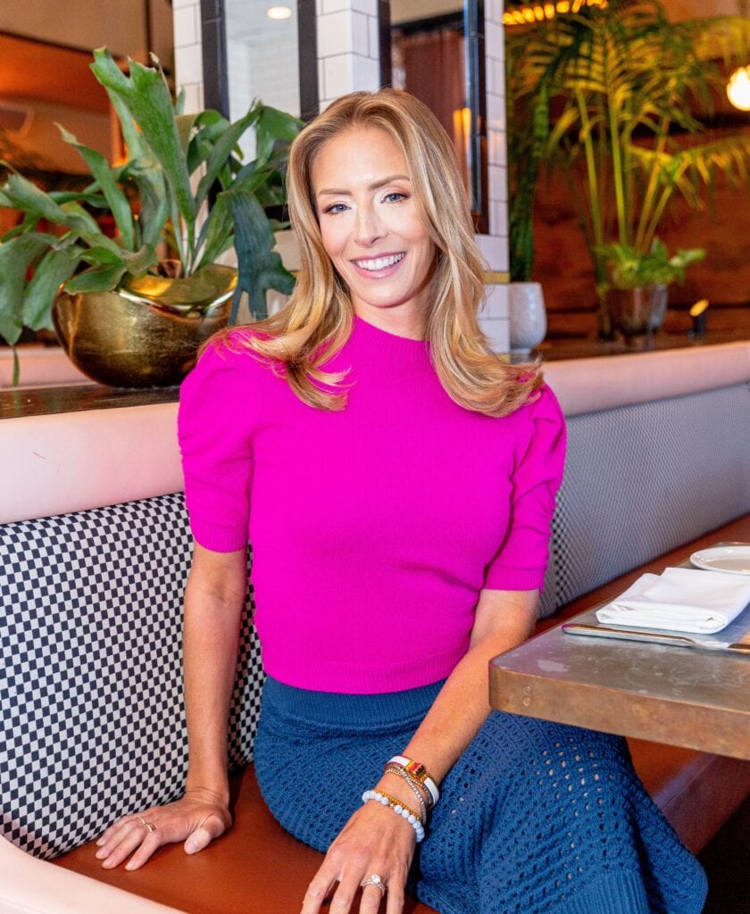 A woman with long blonde hair, wearing a bright pink top and blue skirt, sits smiling at a restaurant booth with a plant and tableware nearby, ready to enjoy an ultimate tailgate-inspired meal.