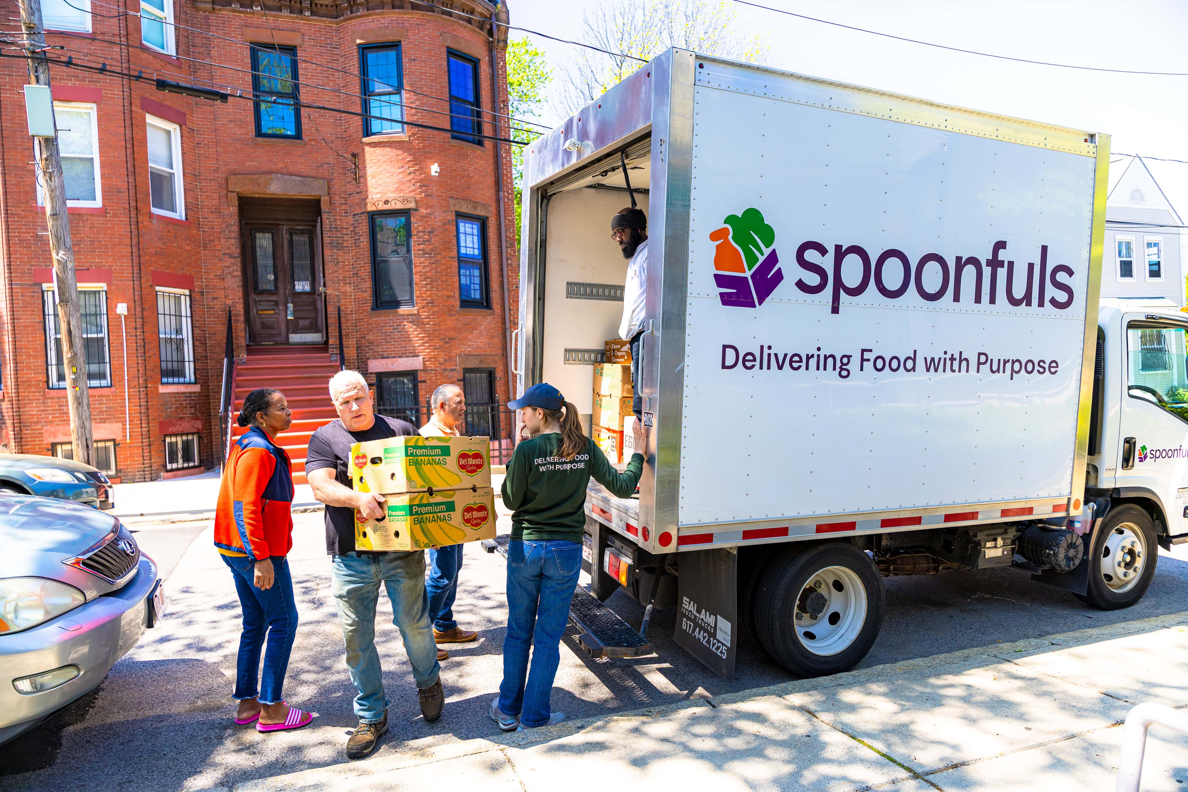 Several people unload boxes of food from a "Spoonfuls" truck on a residential street, participating in a food delivery service.