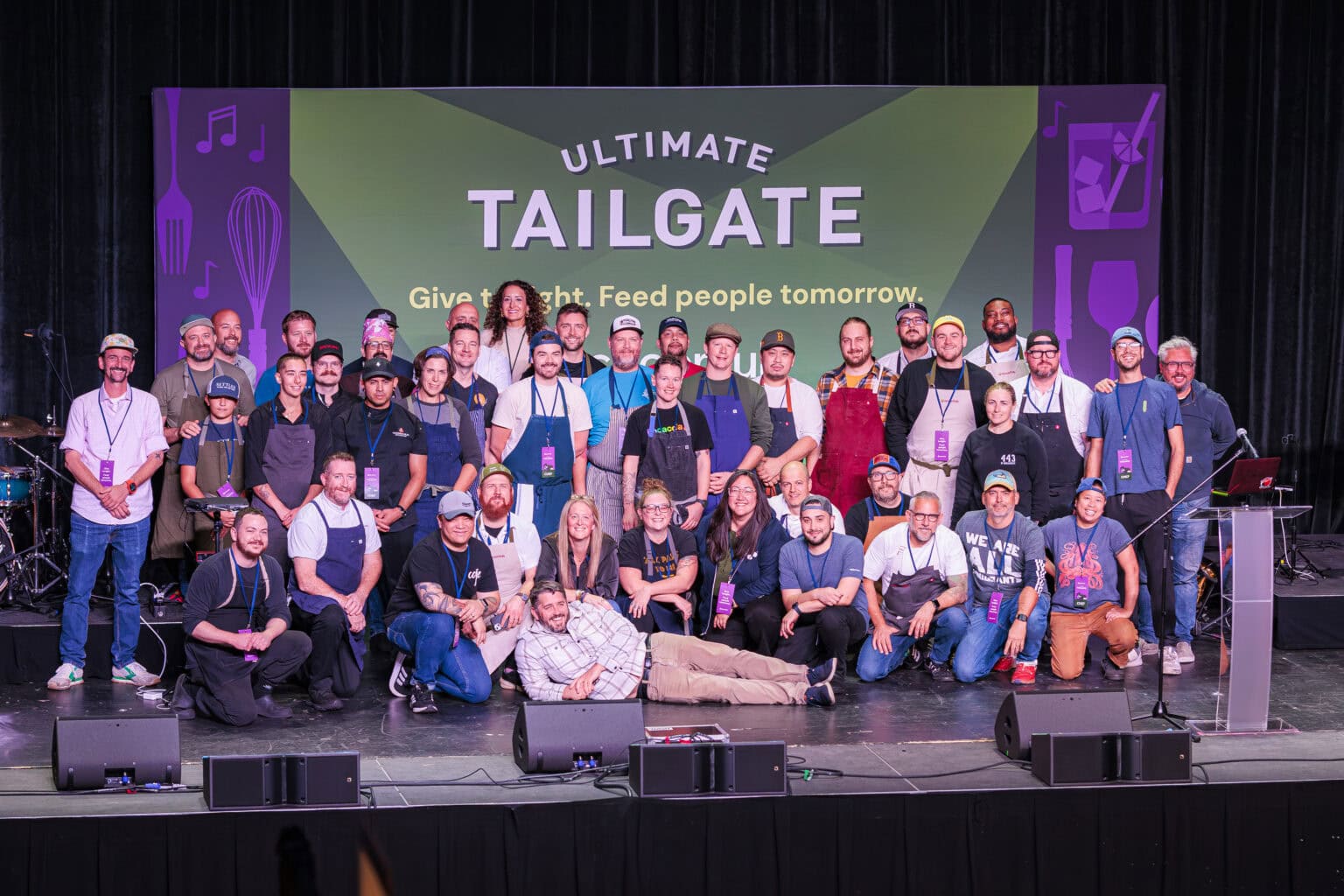A large group of people pose together on stage in front of a green Ultimate Tailgate event banner featuring the slogan "Give a thought. Feed people tomorrow.