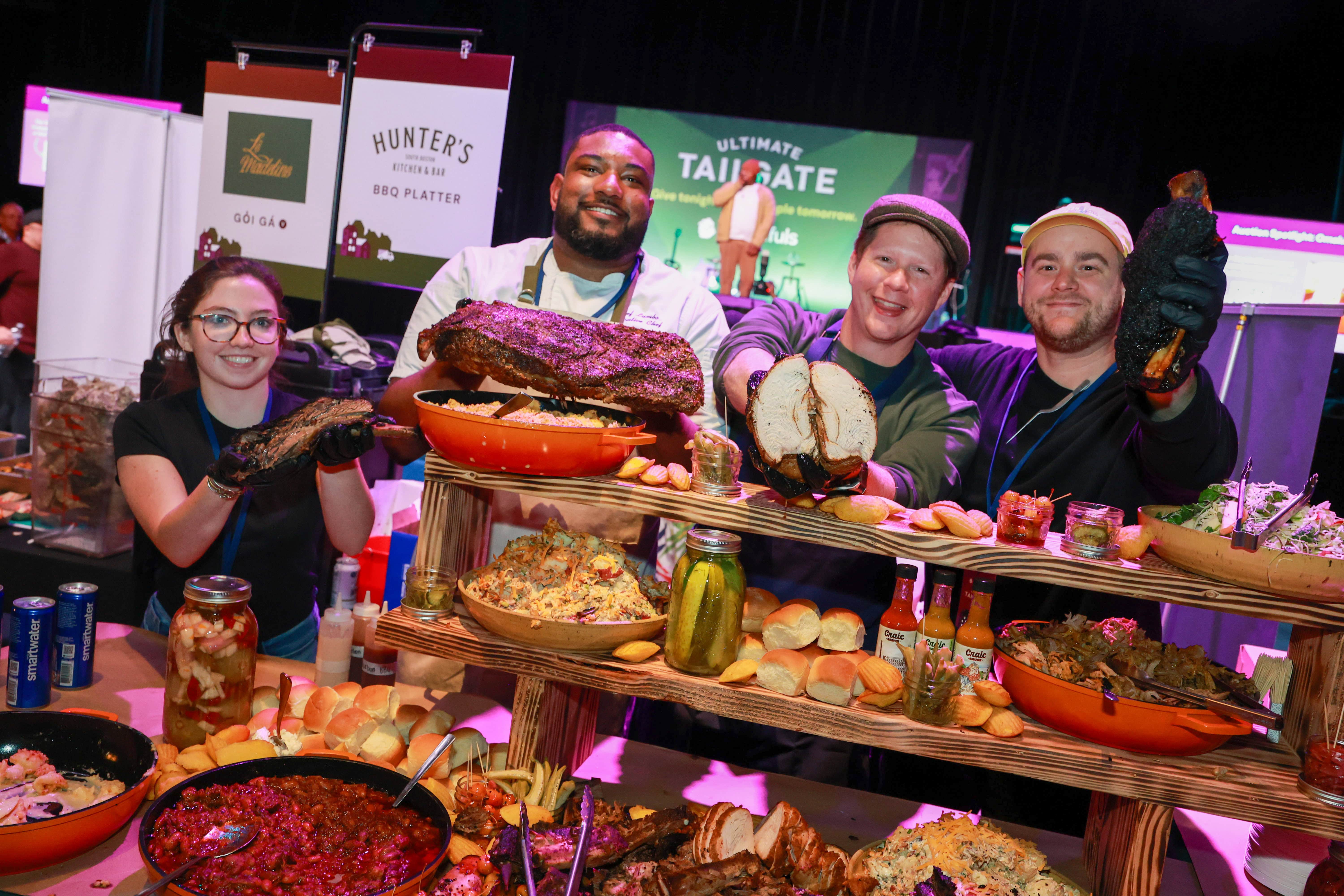 Four people stand behind a table filled with barbecue meats, side dishes, and sliders at the Ultimate Tailgate food event, smiling and holding up samples of food.