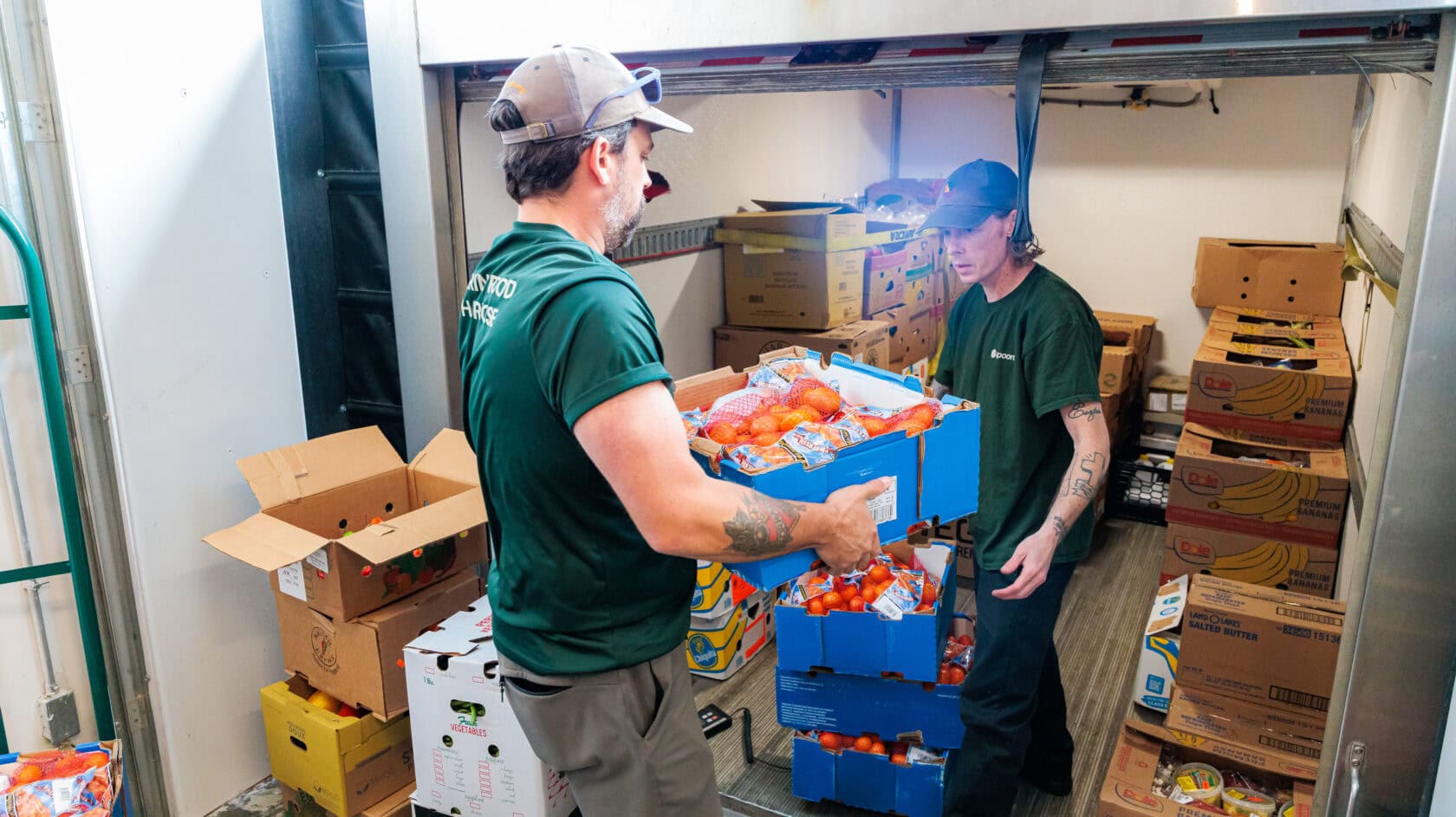 Two workers in green shirts load boxes of packaged produce into a storage area filled with cartons and fresh food items.