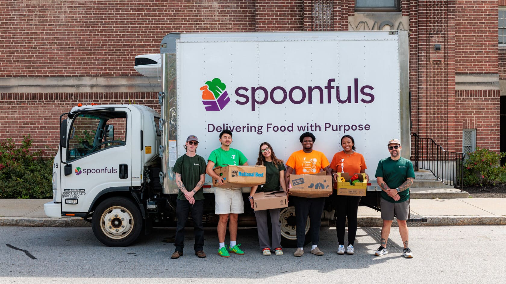 Six people stand in front of a Spoonfuls food truck, each holding boxes of food to donate in Massachusetts, with a brick building in the background.