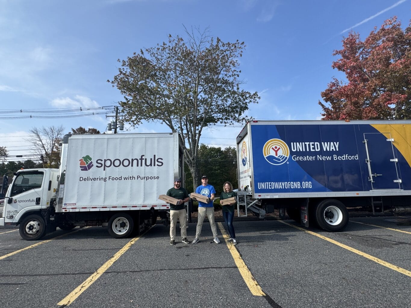 Three people stand in front of Spoonfuls and United Way trucks in a parking lot, each holding a box. The scene is outdoors with trees and clear skies in the background.