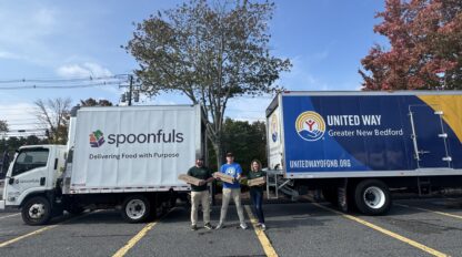 Three people stand in front of Spoonfuls and United Way trucks in a parking lot, each holding a box. The scene is outdoors with trees and clear skies in the background.