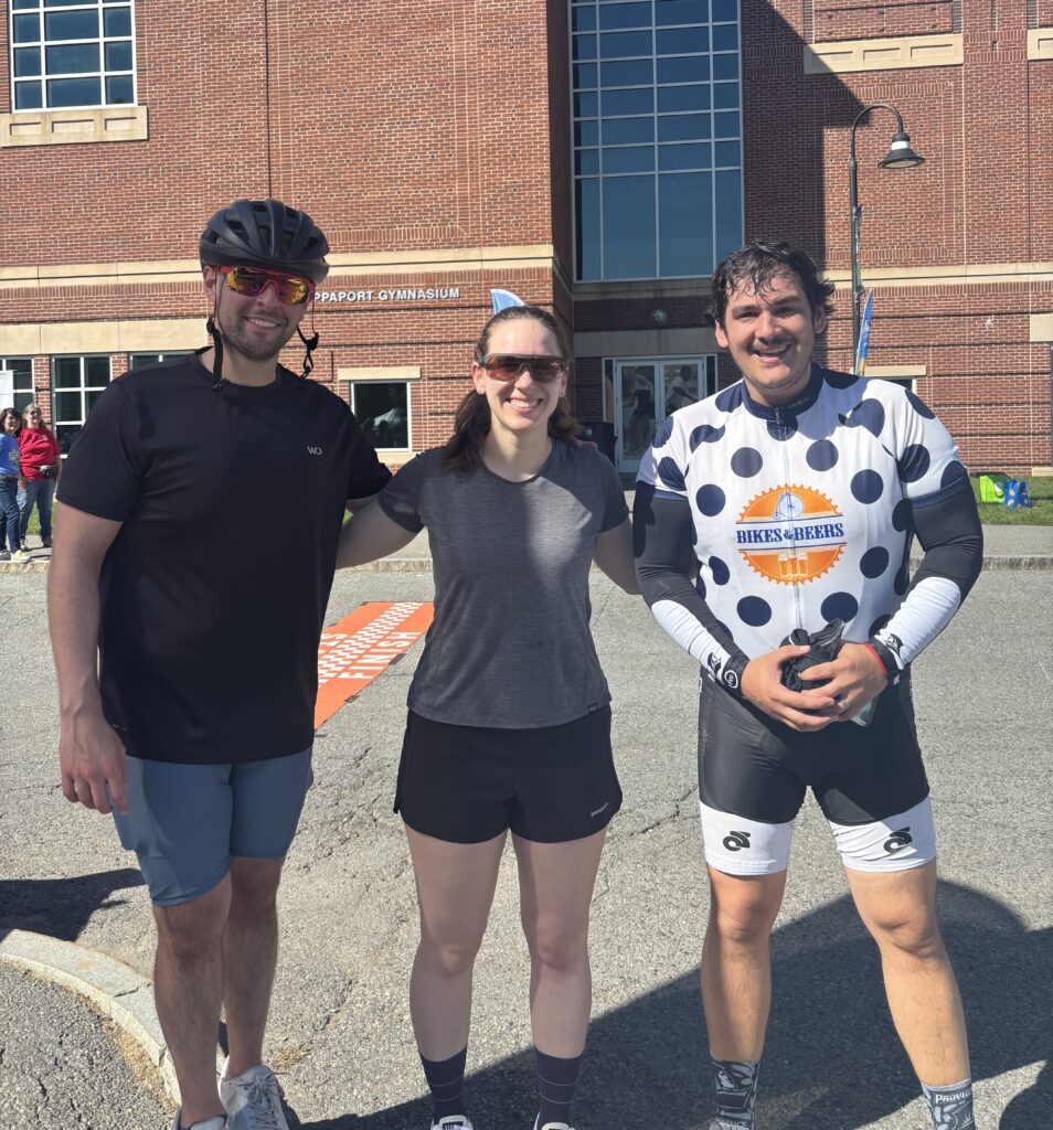 Three people in athletic clothes stand outside a brick building with large windows on a sunny day, ready for the Ride for Food. One person wears a bike helmet, while another sports a cycling jersey.