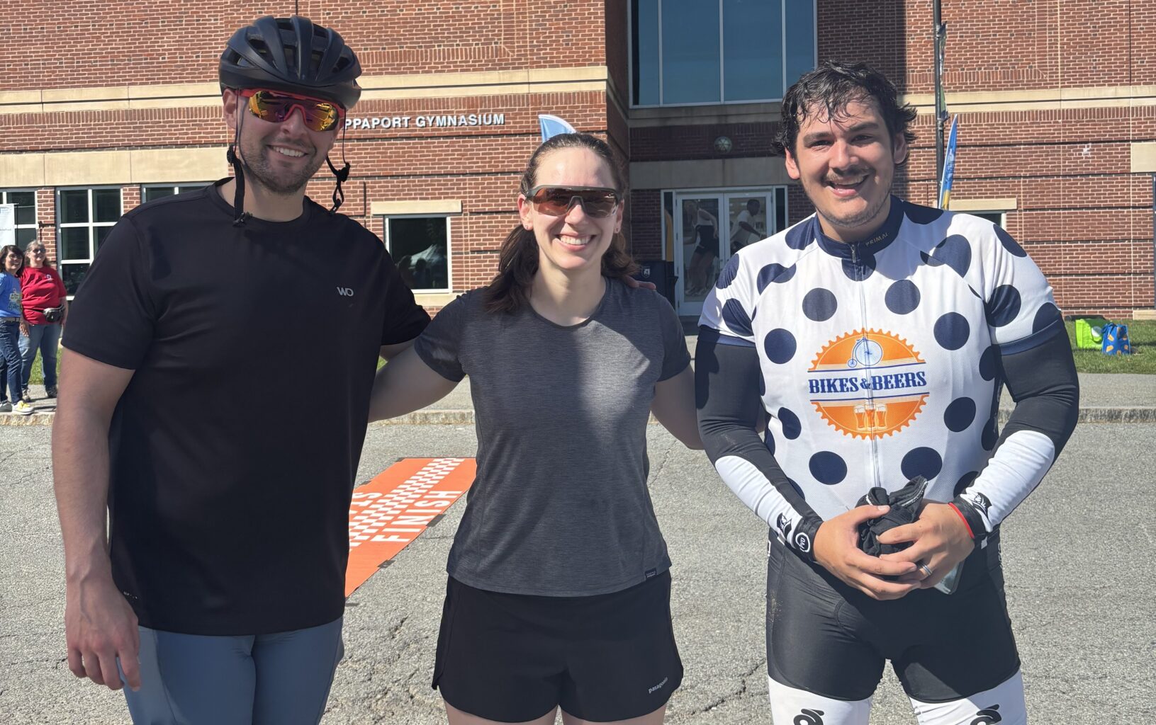 Three people in athletic clothes stand outside a brick building with large windows on a sunny day, ready for the Ride for Food. One person wears a bike helmet, while another sports a cycling jersey.