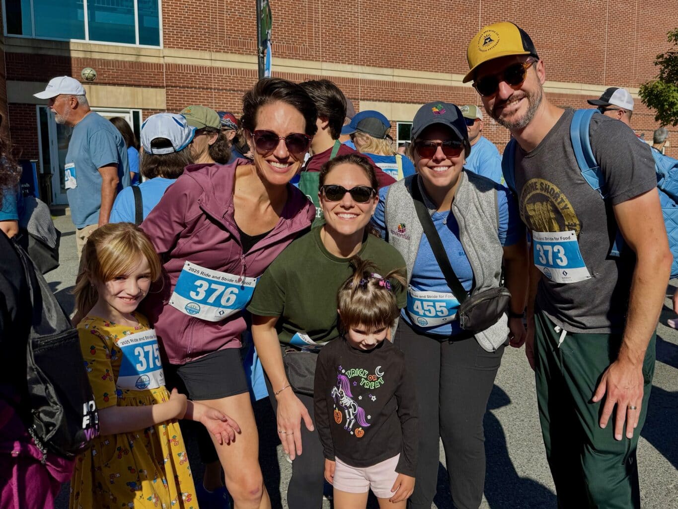 A group of three adults and two children pose outside at the Ride for Food race event, wearing numbered race bibs and casual athletic clothing.