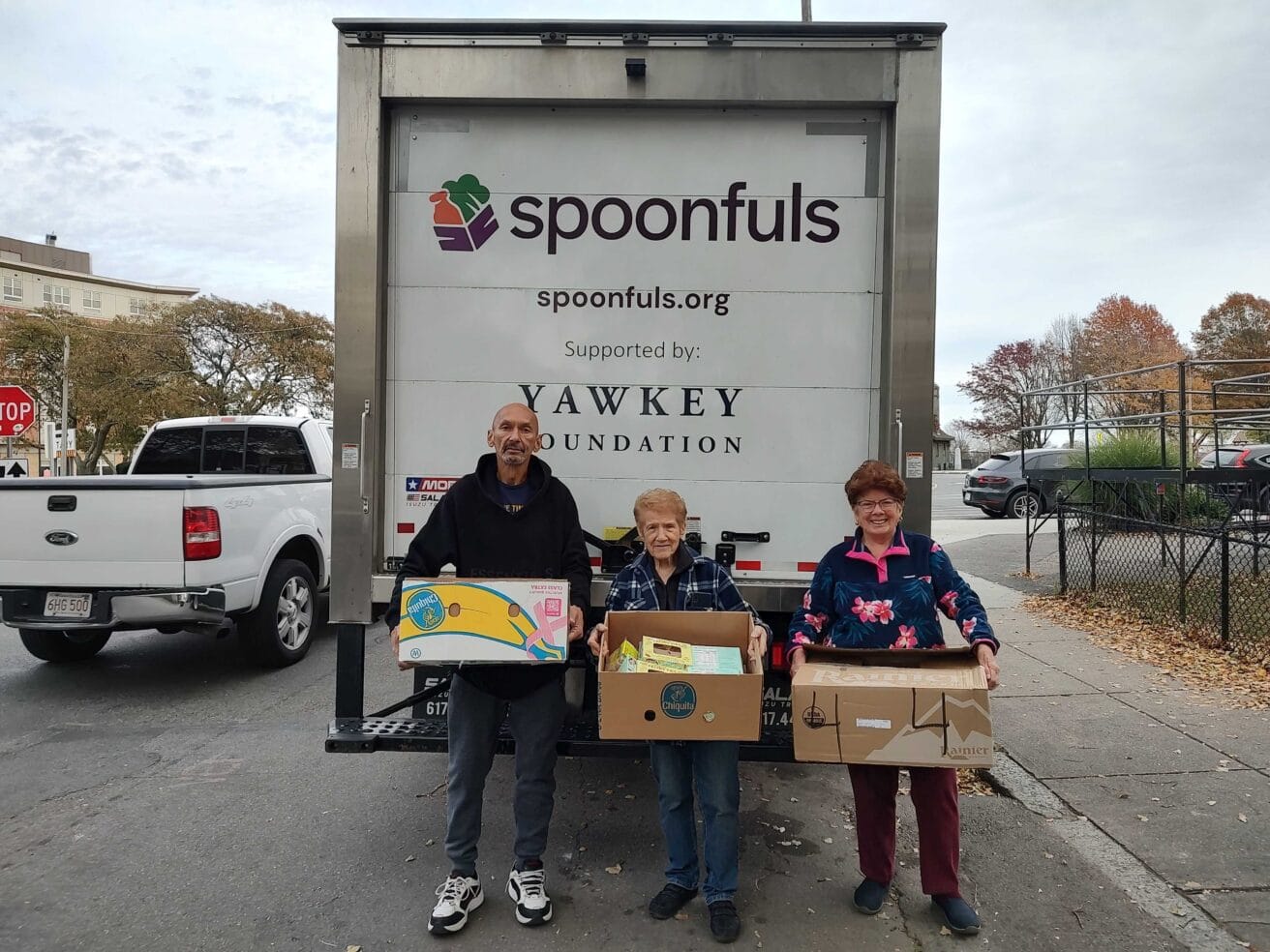 Three people stand in front of a Spoonfuls delivery truck, each holding a box of food items, with buildings and trees in the background.