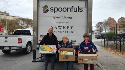 Three people stand in front of a Spoonfuls delivery truck, each holding a box of food items, with buildings and trees in the background.