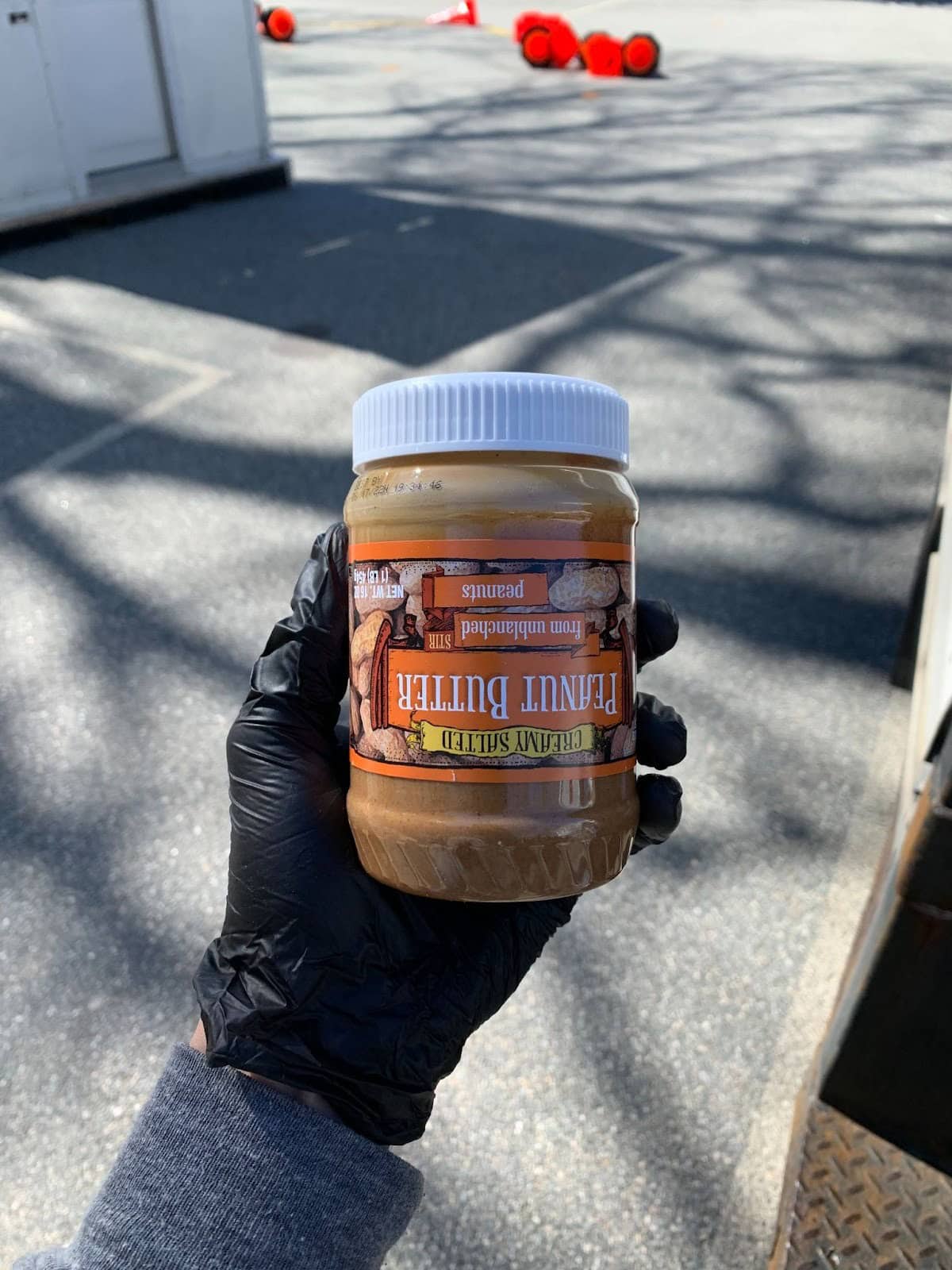 A person wearing a black glove holds a jar of peanut butter labeled "Peanut Butter" outdoors, with red traffic cones visible in the background.