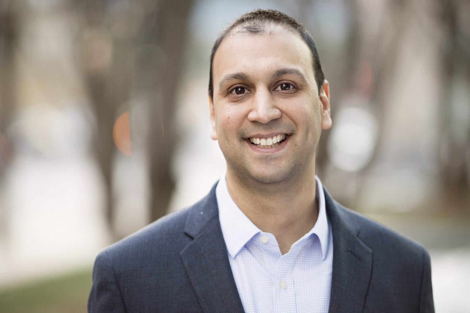 A man in a dark suit jacket and light blue dress shirt smiles at the camera outdoors with a blurred background.