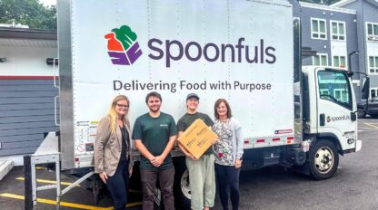 Four people stand in front of a Spoonfuls delivery truck, with one person holding a cardboard box. The truck displays the phrase "Delivering Food with Purpose.