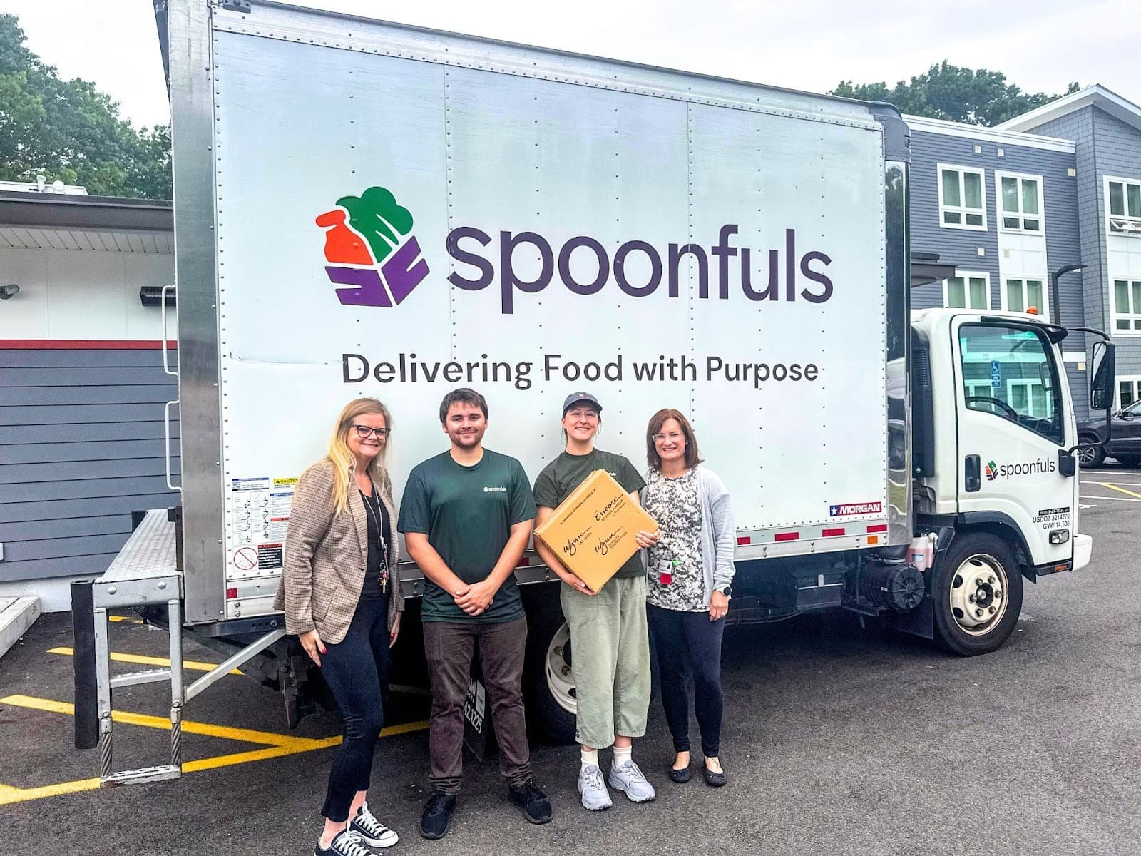 Four people stand in front of a Spoonfuls delivery truck, with one person holding a cardboard box. The truck displays the phrase "Delivering Food with Purpose.