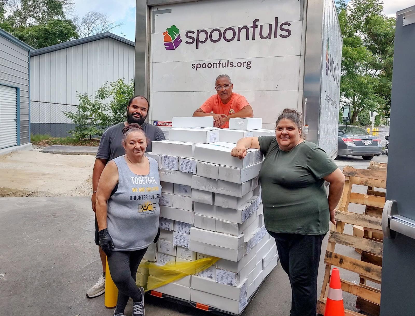 Four people stand around a large stack of boxed goods in front of a Spoonfuls truck outside a warehouse.
