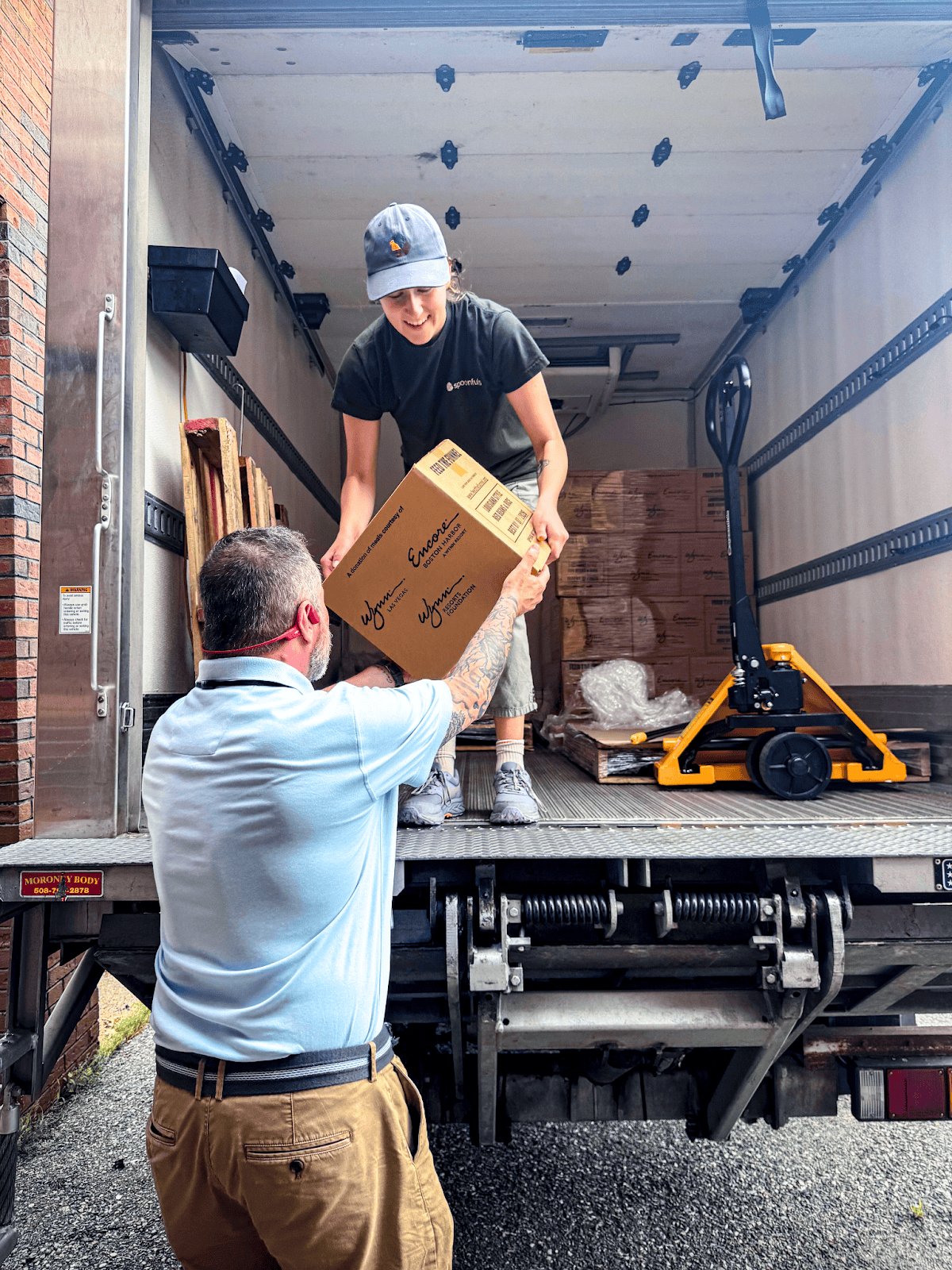 A man stands outside a truck, helping a woman inside the truck unload cardboard boxes onto a pallet jack.