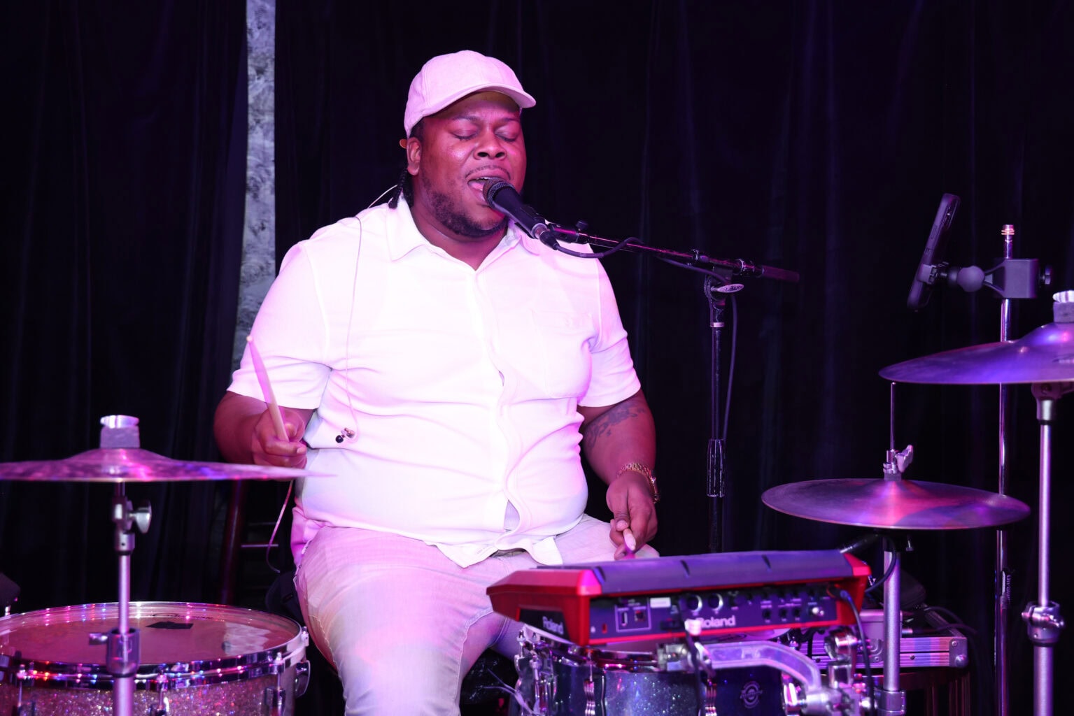 A musician in a white shirt and cap plays drums and sings into a microphone on stage, with a black curtain backdrop, creating the perfect vibe for cocktails & community.