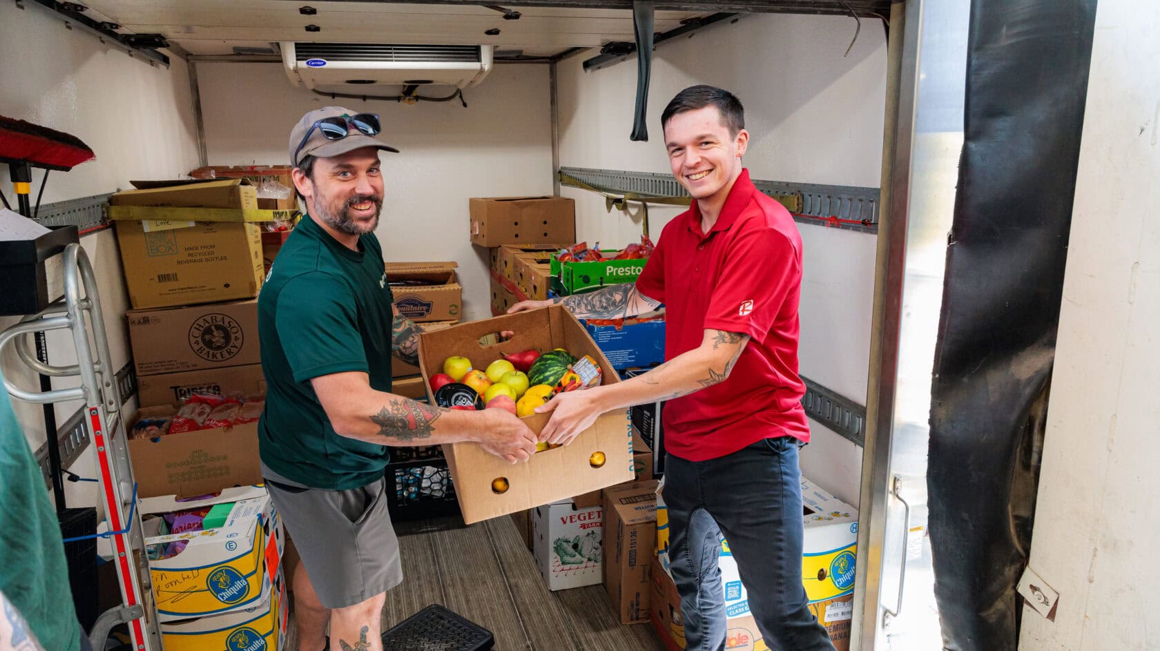 Two men standing in a truck filled with food boxes, holding a box of assorted produce together and smiling at the camera as they help donate food in Massachusetts.