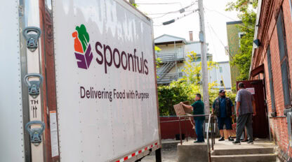 A Spoonfuls truck is parked next to a building as several people stand near a doorway, with one person carrying a box.