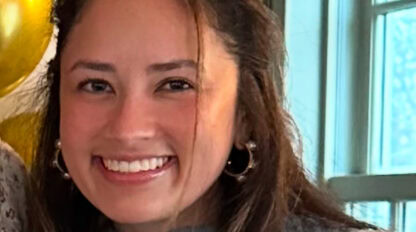A young woman with long brown hair, wearing a grey sweater and hoop earrings, smiles at the camera. Balloons and a window are visible in the background, celebrating after completing the Boston Marathon.
